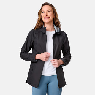 A woman with long brown hair smiles while wearing the Free Country Women's X2O Anorak Rain Jacket in black over a white shirt and light blue pants, standing against a plain light gray background.