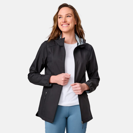 A woman with long brown hair smiles while wearing the Free Country Women's X2O Anorak Rain Jacket in black over a white shirt and light blue pants, standing against a plain light gray background.