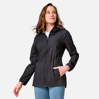 A woman models the Free Country Women's X2O Anorak Rain Jacket—a black, water-resistant hooded jacket with side pockets—paired with light blue pants. She stands against a plain light gray background, smiling and looking to the side.