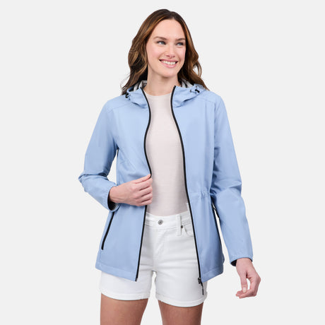 A woman wearing the Free Country Women's X2O Anorak Rain Jacket in light blue over a white shirt and white shorts smiles against a plain white background.