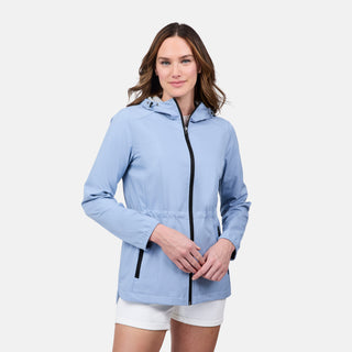A woman with medium-length brown hair wears the Free Country Women's X2O Anorak Rain Jacket in light blue with black accents and a front zipper, paired with white shorts, standing against a plain white background.