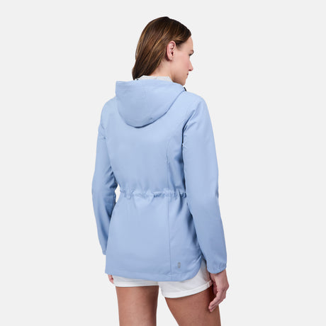 A woman is shown from the back, wearing the Free Country Women's X2O Anorak Rain Jacket in light blue with white shorts, standing against a plain white background.