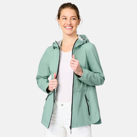 A woman smiles wearing the Free Country Women's X2O Anorak Rain Jacket in light green, unzipped over a white shirt and pants. The jacket features zippered pockets as she stands against a plain light gray background.