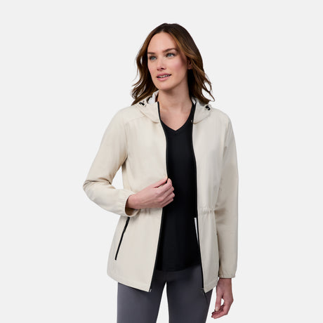 A woman with brown hair wears the Free Country Women's X2O Anorak Rain Jacket in light beige over a black top and gray leggings, standing against a white background and smiling slightly to the side.
