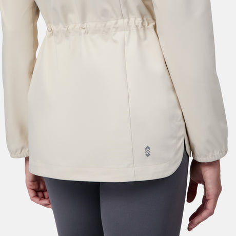 A person is shown from the back wearing the Free Country Women's X2O Anorak Rain Jacket in light beige, featuring an elastic waistband and a small reflective logo by the hem, paired with dark gray leggings.