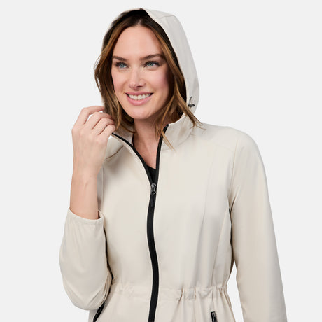 A smiling woman in the Free Country Women's X2O Anorak Rain Jacket, a light beige, water-resistant coat with a front zipper and black accents, holds her hood's edge against a plain white background.