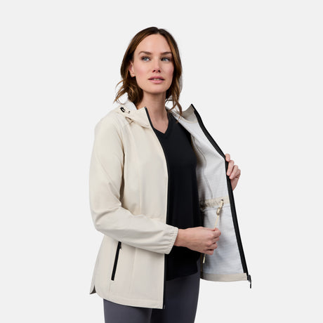 A woman models the Free Country Women's X2O Anorak Rain Jacket in light beige over a black shirt. She stands against a plain background, holding her unzipped jacket open to show the inside pocket.