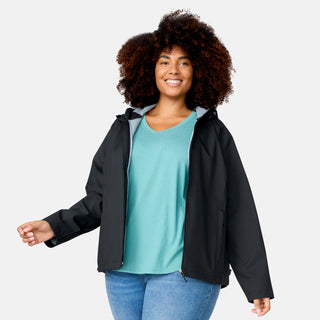 A smiling woman with curly hair wears a teal shirt, blue jeans, and the Free Country Women’s Plus Size X2O Packable Rain Jacket in black. She stands against a plain light background, facing forward with one hand slightly raised.