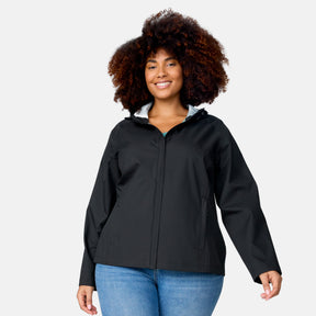 A woman with curly hair wears the Free Country Women's Plus Size X2O Packable Rain Jacket in black and blue jeans, standing against a light gray background and smiling at the camera.