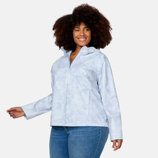 A woman with curly hair wears the Free Country Women's Plus Size X2O Packable Rain Jacket in Silver Chip Floral Bliss and blue jeans, smiling against a plain white background.