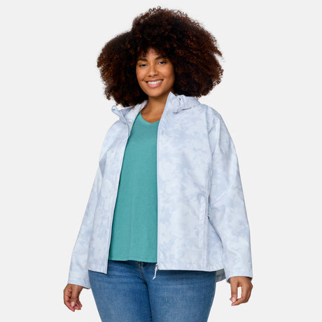 A woman with curly hair smiles, wearing the Free Country Women's Plus Size X2O Packable Rain Jacket in light blue over a teal top and blue jeans, standing against a plain light background.