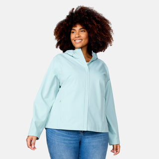 A woman with curly hair is wearing the Free Country Women's Plus Size X2O Packable Rain Jacket in Green Tea and blue jeans. She stands smiling, facing slightly to the side, against a plain light background.