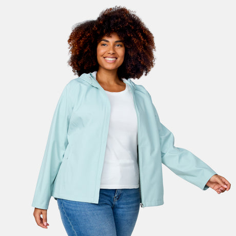 A woman with curly hair smiles, wearing the Free Country Women's Plus Size X2O Packable Rain Jacket in light blue over a white shirt and blue jeans, standing against a plain light background.
