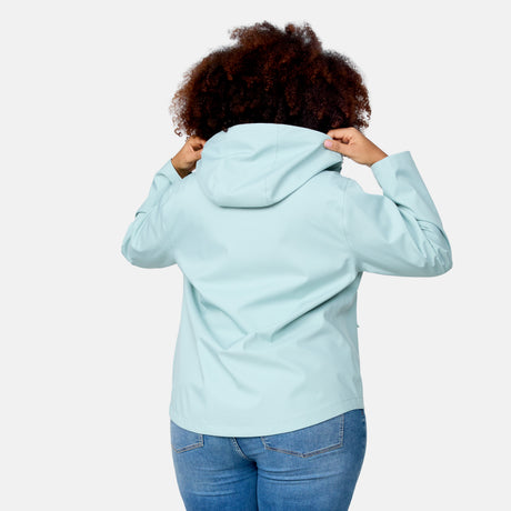 A person with curly hair, seen from behind, wears the Women's Plus Size X2O Packable Rain Jacket by Free Country in light blue and blue jeans, holding up the hood with both hands against a plain white background.