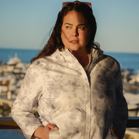 A woman with long dark hair wears the Free Country Women's Plus Size X2O Packable Rain Jacket outdoors in sunlight, hands in her pockets and gazing to the side, with boats and water softly blurred in the background.
