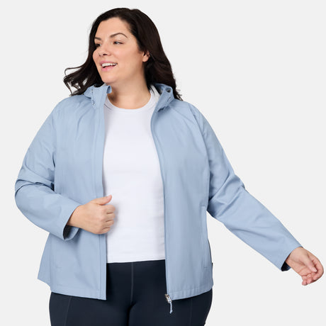 A woman wears the Free Country Women's Plus Size X2O Packable Rain Jacket in light blue over a white shirt and dark pants, standing against a white background, smiling and adjusting her jacket.