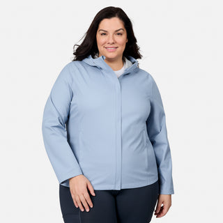 A woman with long dark hair smiles at the camera, wearing the Free Country Women's Plus Size X2O Packable Rain Jacket in light blue and dark pants, standing against a plain white background.