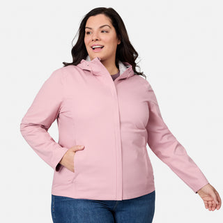 A woman with long dark hair smiles to the side, wearing the Free Country Women's Plus Size X2O Packable Rain Jacket in light pink and blue jeans, one hand in her pocket, against a plain light background.