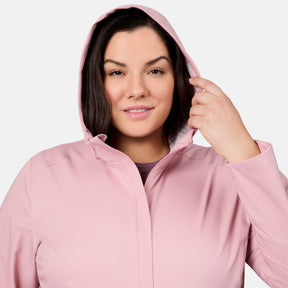A dark-haired woman smiles gently as she adjusts the hood of her light pink Women's Plus Size X2O Packable Rain Jacket by Free Country, set against a plain, light background.