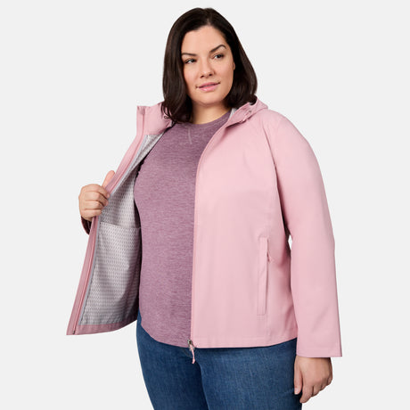 A woman models the Free Country Women's Plus Size X2O Packable Rain Jacket in light pink, wearing it over a mauve shirt and blue jeans, standing against a plain background and holding open the jacket to display its interior lining.