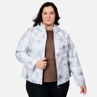 A woman with dark hair models the Free Country Women's Plus Size X2O Packable Rain Jacket in light gray and white over a brown top and black pants, standing against a white background and holding the jacket partially open.