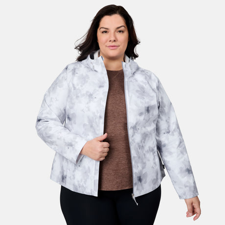 A woman with dark hair models the Free Country Women's Plus Size X2O Packable Rain Jacket in light gray and white over a brown top and black pants, standing against a white background and holding the jacket partially open.