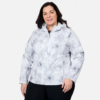 A woman smiles while modeling the Free Country Women's Plus Size X2O Packable Rain Jacket in light gray and white, standing against a plain white background with her left hand at her side and right hand slightly forward.