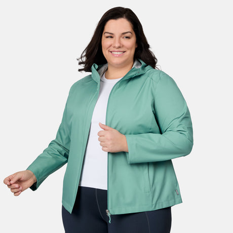 A woman with dark hair smiles while wearing the Free Country Women's Plus Size X2O Packable Rain Jacket in light teal over a white shirt and dark pants, standing against a plain light background.