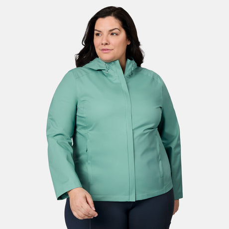 A woman with long dark hair, wearing the Free Country Women's Plus Size X2O Packable Rain Jacket in light teal and dark pants, stands against a plain white background, looking slightly to the side with a neutral expression.