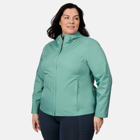 A woman in the Free Country Women's Plus Size X2O Packable Rain Jacket in light teal and dark pants stands slightly turned, smiling against a plain white background.