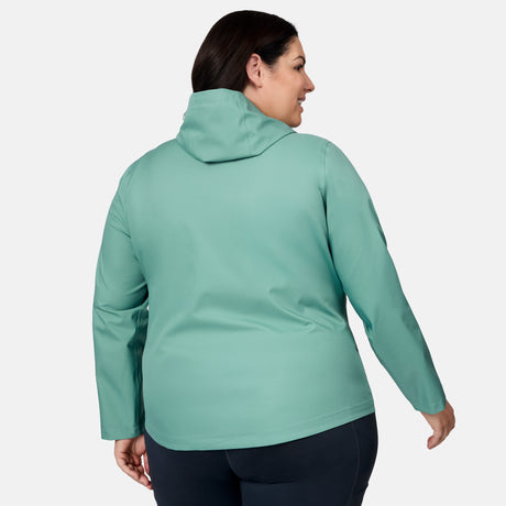 A woman with dark hair is seen from the back, wearing the Free Country Women's Plus Size X2O Packable Rain Jacket in light teal and dark pants, standing against a plain white background.