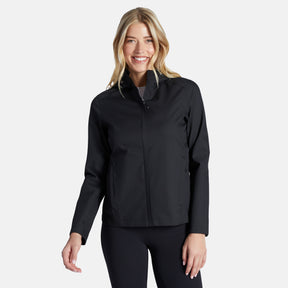 A woman with long blonde hair smiles while wearing the Free Country Women's X2O Packable Rain Jacket in black, paired with matching pants, against a plain light background.