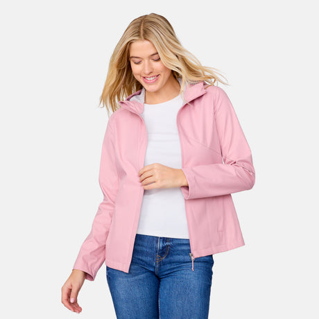 A smiling woman with blonde hair wears the Free Country Women's X2O Packable Rain Jacket in light pink over a white shirt and blue jeans, standing against a plain white background.