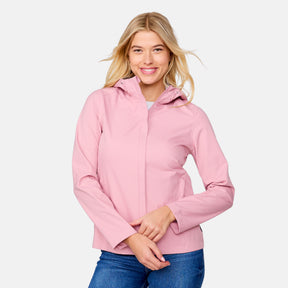 A woman with long blonde hair smiles while wearing the Free Country Women's X2O Packable Rain Jacket in light pink and blue jeans, standing against a plain light gray background.