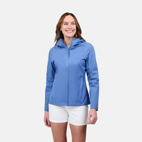 A woman smiles against a white background, wearing the Free Country Women's X2O Packable Rain Jacket in light blue paired with white shorts.