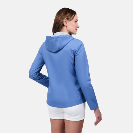 A woman faces away wearing the Free Country Women's X2O Packable Rain Jacket in light blue with white shorts, set against a plain white background.
