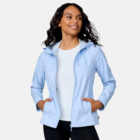 A woman with long, wavy hair smiles and looks to the side while wearing the Free Country Women's X2O Packable Rain Jacket in blue over a white shirt, hands in pockets, standing against a plain light background.