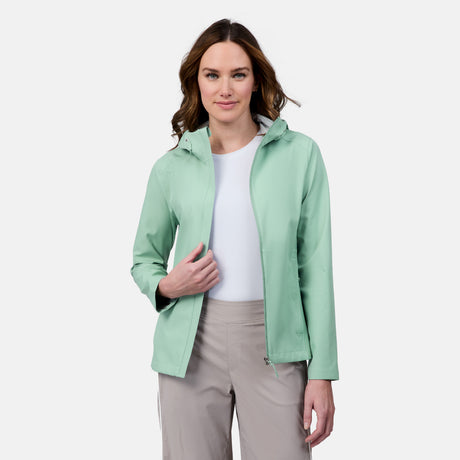 A woman with wavy brown hair models the Free Country Women's X2O Packable Rain Jacket in light green over a white top and beige pants, standing against a plain white background and holding the jacket open with one hand.