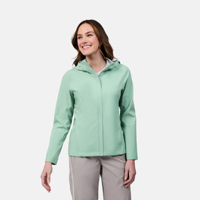 A woman stands smiling against a plain white background, wearing the Free Country Women's X2O Packable Rain Jacket in light green, which is lightweight, breathable, waterproof with UPF 50+ protection, and light gray pants.