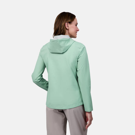 A woman faces away, wearing the Free Country Women's X2O Packable Rain Jacket in light green with light-colored pants, set against a plain white background.