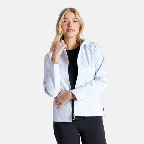 A woman with long blonde hair wears the Free Country Women's X2O Packable Rain Jacket—lightweight, breathable, and UPF 50+—over a black top and pants, standing against a plain white background and smiling softly.