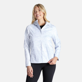 A woman with long blonde hair smiles while wearing the Free Country Women's X2O Packable Rain Jacket in light gray over a black top and leggings, standing against a plain white background.