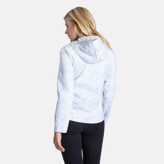 A woman with blonde hair, wearing the Free Country Women's X2O Packable Rain Jacket and black leggings, stands facing away from the camera against a plain white background.