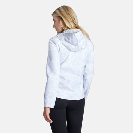 A woman with blonde hair, wearing the Free Country Women's X2O Packable Rain Jacket and black leggings, stands facing away from the camera against a plain white background.