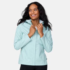 A woman with long, wavy black hair smiles while wearing the Free Country Women's X2O Packable Rain Jacket in light blue, holding the collar with both hands, standing against a plain white background.