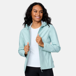 A woman with long, wavy dark hair smiles while wearing the Free Country Women's X2O Packable Rain Jacket in light blue over a white shirt and black pants, standing against a plain light background.