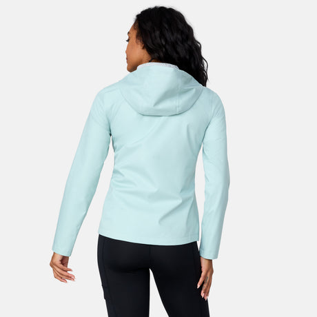 A woman with long wavy hair stands against a light background, facing away to show the back of the Free Country Women's X2O Packable Rain Jacket, which is lightweight and breathable. She wears black leggings.