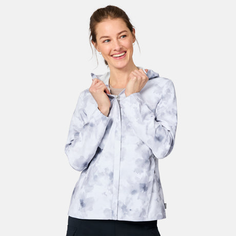 A smiling woman stands against a plain background, wearing the Free Country Women's X2O Packable Rain Jacket in gray and white tie-dye with the hood up, her hands holding the front near her neck.