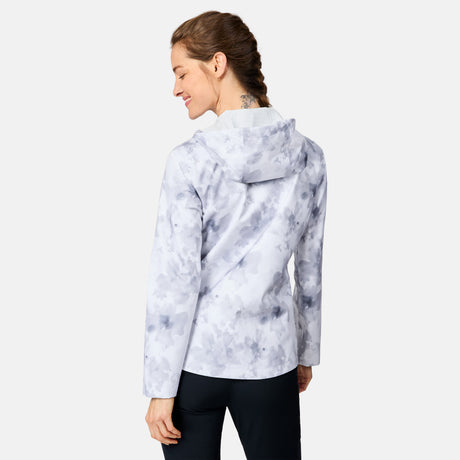 A woman smiles over her shoulder, wearing the Free Country Women's X2O Packable Rain Jacket—lightweight, breathable, and UPF 50+—with black pants, against a plain light background.