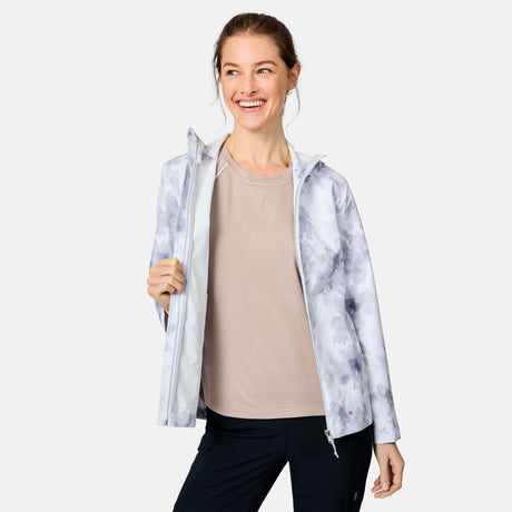 A woman smiles while wearing the Free Country Women's X2O Packable Rain Jacket in light beige with UPF 50+ protection, holding it open against a plain white background.